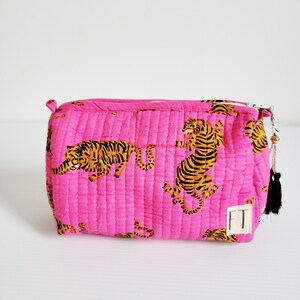 Op de afbeelding: Een gewatteerde, rechthoekige make-up tas in felroze, met een herhalend patroon van oranje en zwarte tijger illustraties. De tas heeft een ritssluiting en een kleine zwarte kwast met gouden kralen. Een klein wit label met de letters "FT" is aan de zijkant genaaid.