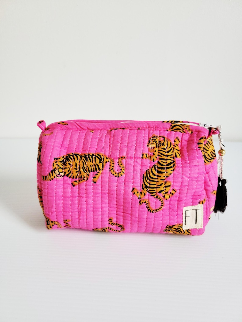 Op de afbeelding: Een gewatteerde, rechthoekige make-up tas in felroze, met een herhalend patroon van oranje en zwarte tijger illustraties. De tas heeft een ritssluiting en een kleine zwarte kwast met gouden kralen. Een klein wit label met de letters "FT" is aan de zijkant genaaid.