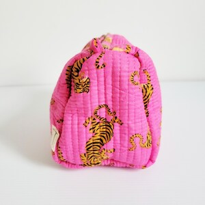 Op de afbeelding: Een gewatteerde, roze make-up tas met een herhalend patroon van oranje en zwarte tijger illustraties. De tas heeft een rechthoekige vorm met afgeronde randen en verticale stiksels. Een klein wit label is zichtbaar.