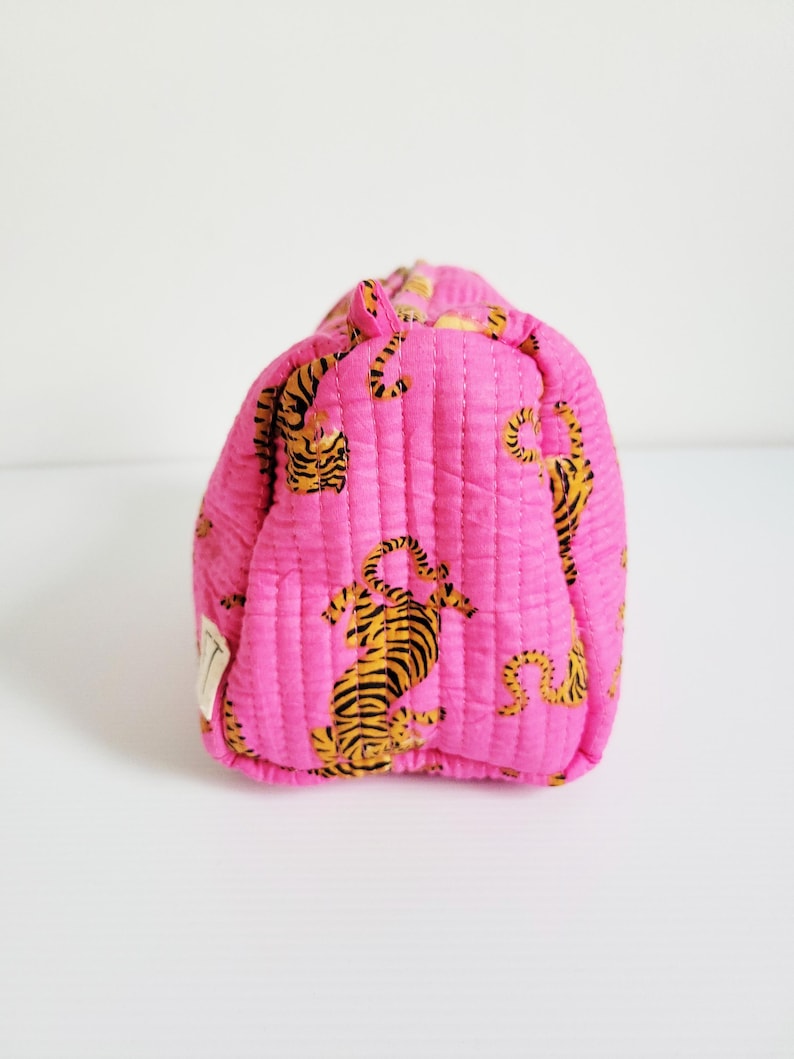 Op de afbeelding: Een gewatteerde, roze make-up tas met een herhalend patroon van oranje en zwarte tijger illustraties. De tas heeft een rechthoekige vorm met afgeronde randen en verticale stiksels. Een klein wit label is zichtbaar.
