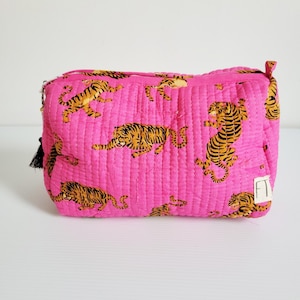 Op de afbeelding: Een gewatteerde, rechthoekige make-uptas in een levendige roze tint, versierd met een herhalend patroon van oranje en zwarte tijgerillustraties. De tas heeft een zwarte kwastjesritssluiting en een klein wit label met zwarte tekst.