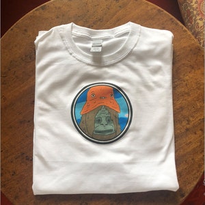 Peut inclure: T-shirt blanc avec un graphique circulaire d'une créature verte portant un chapeau orange. La créature a une expression surprise et est entourée d'un fond bleu.