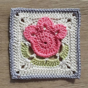 Puede incluir: Un cuadrado tejido a crochet con una flor rosa en el centro. La flor tiene cinco pétalos y está rodeada de hojas verdes. El cuadrado está bordeado con un borde de crochet gris.