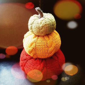 May include: A stack of three knitted pumpkins in red, yellow, and green. The pumpkins are made with a cable knit pattern.