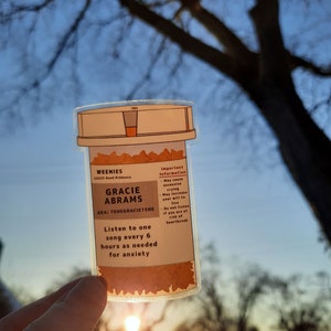 May include: A clear plastic pill bottle with a brown label that reads "WEENIES 22423 Good Riddance GRACIE ABRAMS AKA: TEHEGRACIETEHE Listen to one song every 6 hours as needed for anxiety Important Information - May cause excessive crying - May increase your will to live Do not listen if you are at risk of heartbreak"