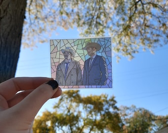 May include: A stained glass style sticker featuring two men in suits and hats. The men are standing side by side against a blue background. The sticker is held in a hand against a blurred background of trees and a house.