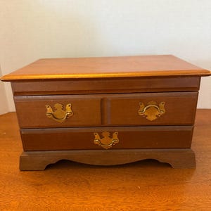 May include: A rectangular wooden jewelry box with a medium brown finish. The box features two drawers, each equipped with decorative brass-colored metal pulls. The base of the box has a scalloped edge design, and the top surface is flat.