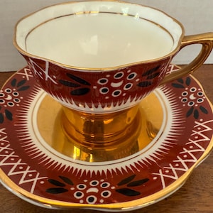 May include: A porcelain teacup and saucer set featuring a deep red background with black leaf motifs and white circular patterns. The cup has a gold-toned handle and base, while the saucer features a matching red, black, and white design.