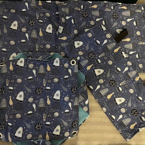 May include: Four blue fabric squares with a white and grey space-themed pattern. The pattern includes rockets, stars, planets, and comets. The squares are different sizes and have gold eyelets in the corners.