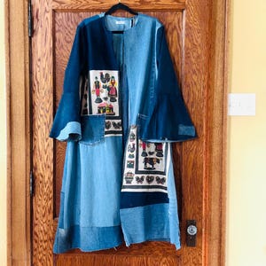 May include: A patchwork denim duster coat in shades of blue. The coat features bell sleeves and decorative panels with colorful folk art motifs. The coat is hanging on a black hanger against a wooden door.