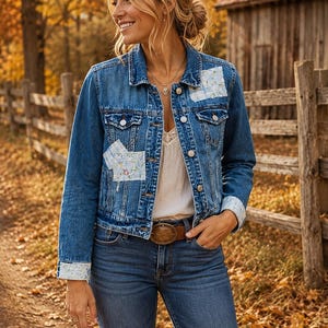 May include: A blue denim jacket featuring light-colored floral patterned fabric patches on the chest and cuffs. The jacket has a classic collar, metal button closures, and two chest pockets. It is worn over a white top with blue denim jeans.