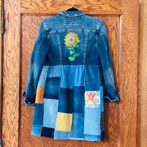 May include: A denim jacket with a patchwork skirt. The back of the jacket features a sunflower embroidery. The skirt is made of various denim patches with colorful accents. The jacket is hanging on a black hanger.