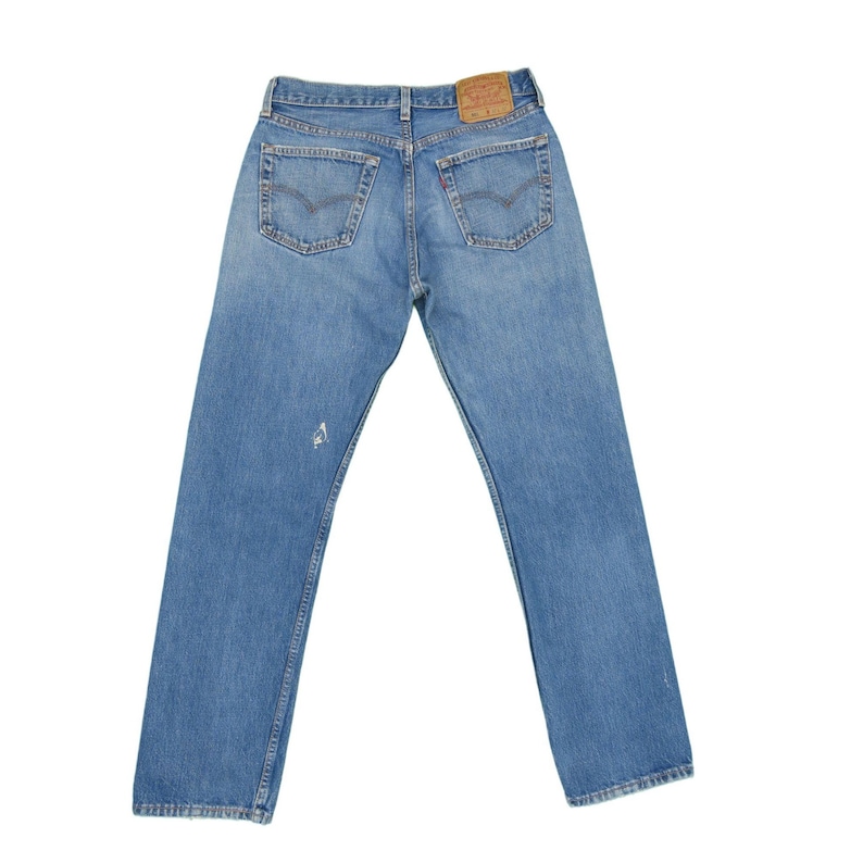 May include: A pair of blue denim jeans with a brown leather patch on the back waistband. The jeans have a faded look and a small hole on the left leg.