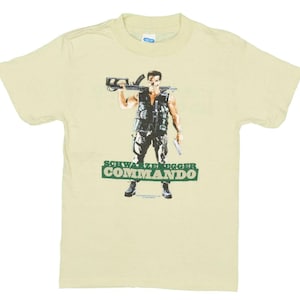 Puede incluir: Una camiseta de color amarillo claro con un estampado gráfico de Arnold Schwarzenegger como John Matrix de la película Commando. El gráfico lo muestra sosteniendo un rifle y una pistola. El texto "Schwarzenegger Commando" está impreso debajo del gráfico.