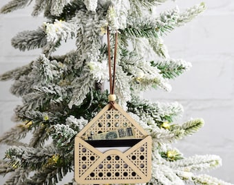 Puede incluir: Un adorno navideño de madera con forma de sobre con un patrón tejido. El adorno tiene un cordón de cuero marrón y está colgado de un árbol de Navidad artificial nevado.