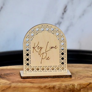May include: Wooden sign with a decorative arch design and the text "Key Lime Pie" in a cursive font.