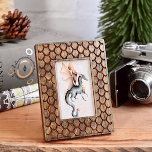 May include: A wooden picture frame with a honeycomb pattern, featuring a watercolor painting of a green dragon with orange wings. The frame is sitting on a wooden surface.