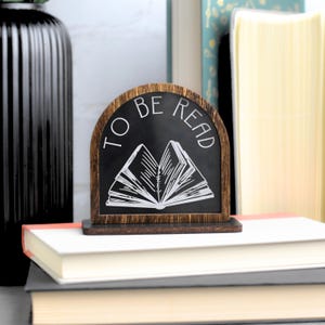 May include: A wooden sign with a black chalkboard insert that reads "To Be Read" in white text. The chalkboard has a white illustration of an open book.