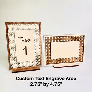 Op de afbeelding: Twee houten tafelnummerhouders met een geweven ontwerp. De grotere houder heeft de tekst "Tafel 1" gegraveerd. De kleinere houder is leeg. De tekst "Aangepaste tekstgraveergebied 2,75" x 4,75" is onder de houders gedrukt.