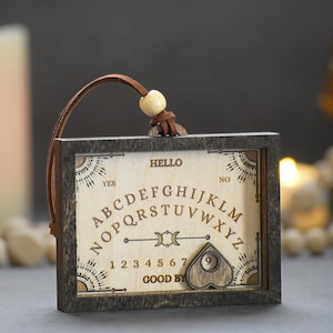 May include: A wooden Ouija board with a dark frame and a light-colored surface. The board features the alphabet, numbers, and the words "HELLO" and "GOOD BYE". A wooden planchette and a leather strap are attached.
