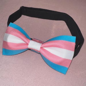 May include: A bow tie featuring horizontal stripes in light blue, pink, and white, arranged in the pattern of the transgender pride flag. The bow tie is attached to a solid black adjustable neck strap with a metal slider for secure fastening.