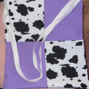 May include: A tote bag featuring a four-panel patchwork design with alternating sections of white fabric with black cow print spots and solid light purple fabric. The bag includes two long white fabric shoulder straps attached at the top.