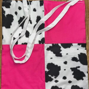 May include: A square tote bag featuring a four-panel patchwork design. Two panels display a black and white cow print pattern, while the other two panels are solid hot pink. The bag includes two long white fabric shoulder straps attached at the top.
