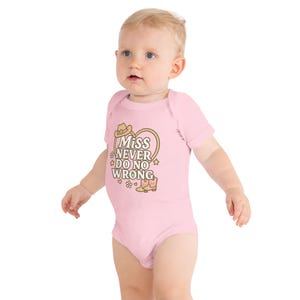 May include: A light pink baby onesie with short sleeves. The onesie features a graphic with a cowboy hat, heart, stars, and boots, along with the text "Miss Never Do No Wrong" in a playful font.