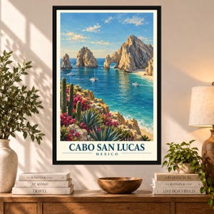 May include: A framed vertical travel poster featuring a scenic view of Cabo San Lucas, Mexico. The artwork depicts rocky coastal cliffs, turquoise ocean waters with small boats, and foreground cacti with pink flowers. The text reads CABO SAN LUCAS MEXICO.