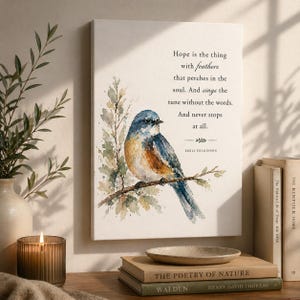 May include: A canvas print featuring a watercolor painting of a bluebird perched on a branch. The artwork includes the quote "Hope is the thing with feathers" by Emily Dickinson. The print is displayed on a wooden surface with books and a candle.