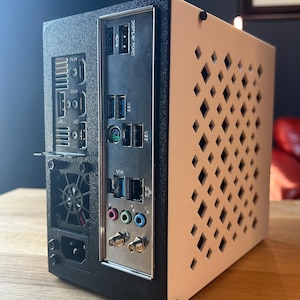 May include: A black and white computer case with a diamond pattern on the side. The back panel has various ports for display, USB, and audio. The case is on a wooden surface.