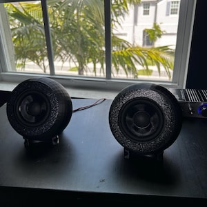 Peut inclure: Deux enceintes sphériques noires avec un bord texturé et scintillant sont posées sur une surface sombre. Les enceintes sont placées devant une fenêtre avec vue sur des palmiers et une maison. Un fil relie les enceintes.