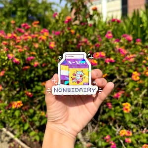 May include: A sticker featuring a cartoon milk carton design with a cow illustration. The carton is purple, yellow, and white, with Japanese text and the words "NONBIDAIRY" below. The sticker is held in a hand against a background of colorful flowers.