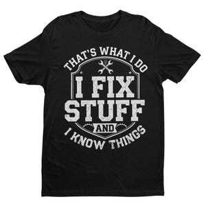 Op de afbeelding: Zwart T-shirt met witte tekst: "THAT'S WHAT I DO I FIX STUFF AND I KNOW THINGS." Het ontwerp bevat gekruiste sleutels en een schildvorm. Het T-shirt heeft een ronde hals.