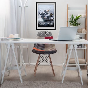 Op de afbeelding: Een wit bureau met een witte stoel en een laptop. Aan de muur achter het bureau hangt een ingelijste poster van de videogame "God of War Ragnarok". De poster toont een sc&egrave;ne waarin Kratos en Atreus vechten tegen een gigantische wolf.