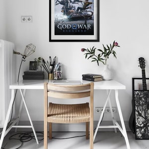 Op de afbeelding: Een zwart ingelijste poster met een blauwe en witte illustratie van drie figuren op een slee. De poster heeft de tekst "God of War Ragnar&ouml;k" in witte letters.