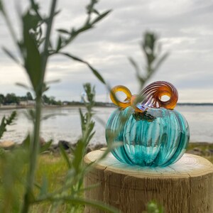 May include: A small, hand-blown glass pumpkin sculpture featuring a translucent teal, ribbed body and a spiraled, multi-colored stem in shades of amber and brown. The glass object sits atop a cylindrical wooden post with visible grain patterns.