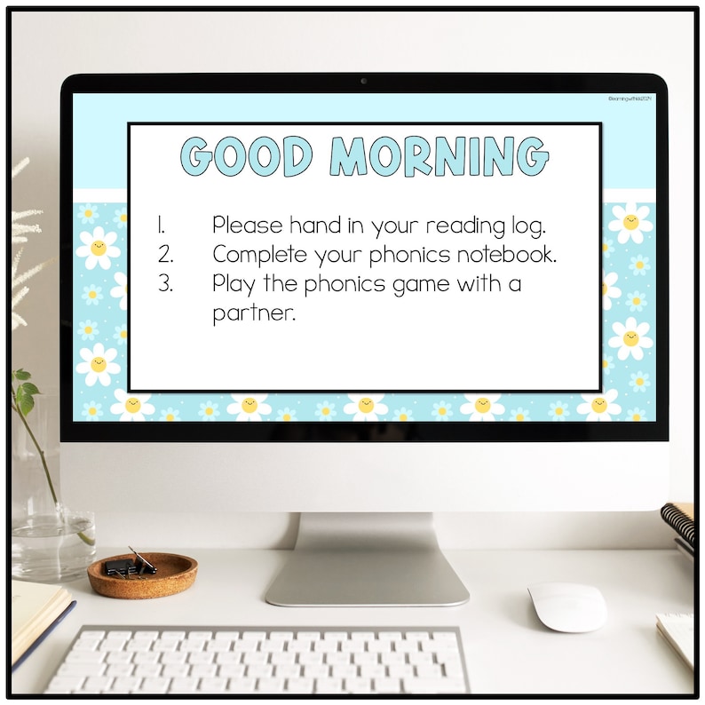 May include: A computer screen displaying a blue and white background with a daisy pattern. The screen shows a list of morning classroom instructions: "GOOD MORNING 1. Please hand in your reading log. 2. Complete your phonics notebook. 3. Play the phonics game with a partner."