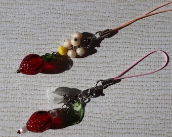 May include: Two red glass strawberry charms with green leaves and white beads on pink and white string phone charms.
