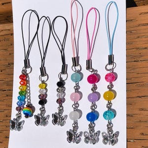 May include: A collection of colorful beaded phone charms. Each charm features a silver butterfly charm and a variety of beads in colors like rainbow, yellow, pink, and blue. The charms have black, pink, and blue cords.