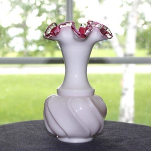 May include: A white milk glass vase featuring a bulbous base with a swirling ribbed pattern and a narrow neck. The top rim is flared and ruffled, accented with a vibrant pink edge that contrasts against the opaque white body of the glass vessel.