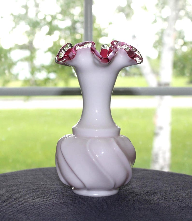 May include: A white milk glass vase featuring a bulbous base with a swirling ribbed pattern and a narrow neck. The top rim is flared and ruffled, accented with a vibrant pink edge that contrasts against the opaque white body of the glass vessel.