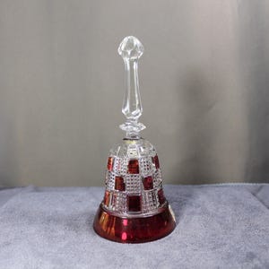 May include: A clear crystal bell with a ruby red base and accents. The bell has a long, clear handle and a checkered pattern of clear and red squares. The bell is likely made of cut crystal.