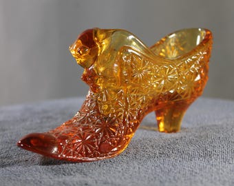May include: An amber-colored glass shoe figurine with a starburst pattern. The decorative shoe has a pointed toe, a high heel, and a detailed design. The glass is transparent, allowing light to pass through.