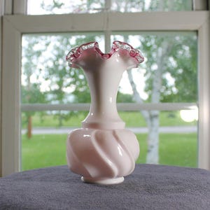 May include: A pale pink glass vase with a bulbous, spiraled base and a slender neck. The rim features a ruffled edge with a deep pink trim. The glass has an opaque, milk-like finish, creating a smooth appearance throughout the body of the vessel.