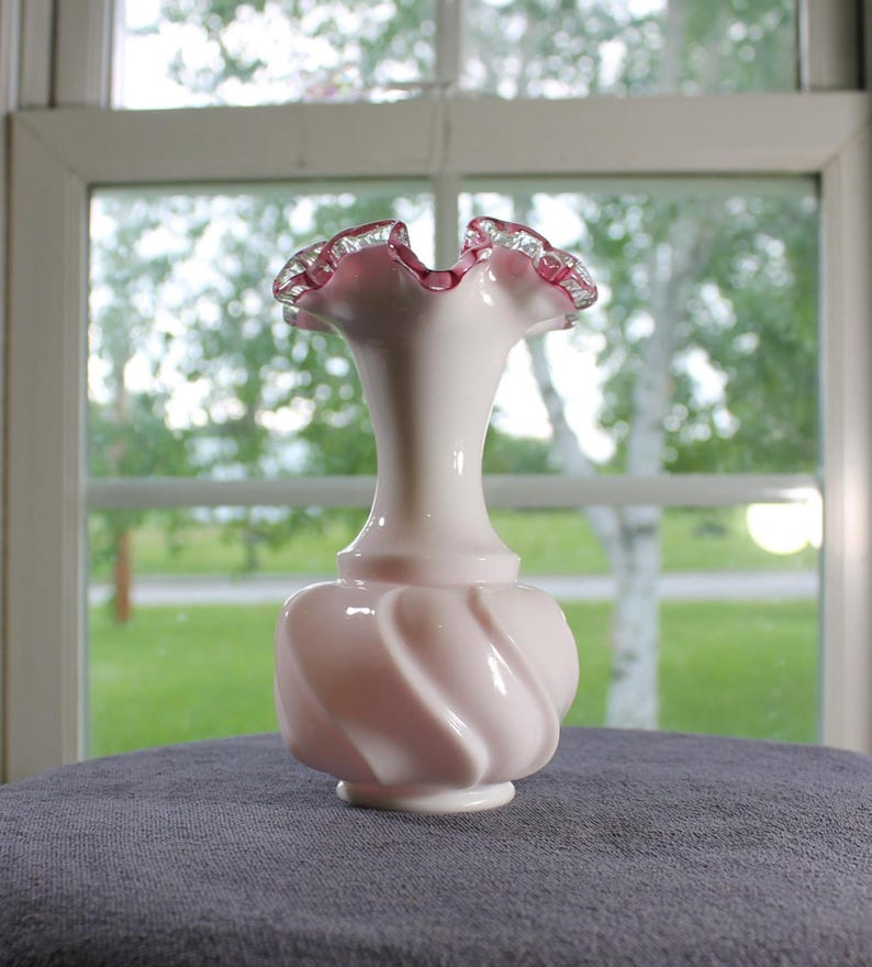 May include: A pale pink glass vase with a bulbous, spiraled base and a slender neck. The rim features a ruffled edge with a deep pink trim. The glass has an opaque, milk-like finish, creating a smooth appearance throughout the body of the vessel.