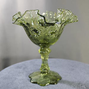 May include: A green glass pedestal bowl with a ruffled rim. The bowl has a floral pattern and sits on a stem with a leaf-shaped base. The glass has a slight yellow tint.