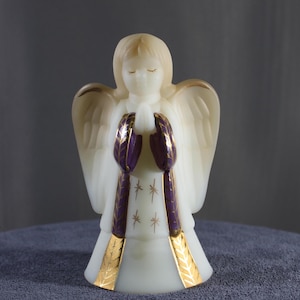 May include: A cream-colored angel figurine with closed eyes and folded hands. The angel's wings and halo have gold accents. The purple and gold details on the angel's sleeves and dress add a touch of elegance. The dress features gold starburst designs.