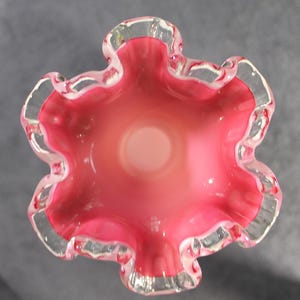 May include: A top-down view of a glass vase featuring a vibrant pink interior that transitions to a lighter, translucent center. The rim is characterized by a distinct, ruffled, and wavy edge with a clear, transparent glass border.