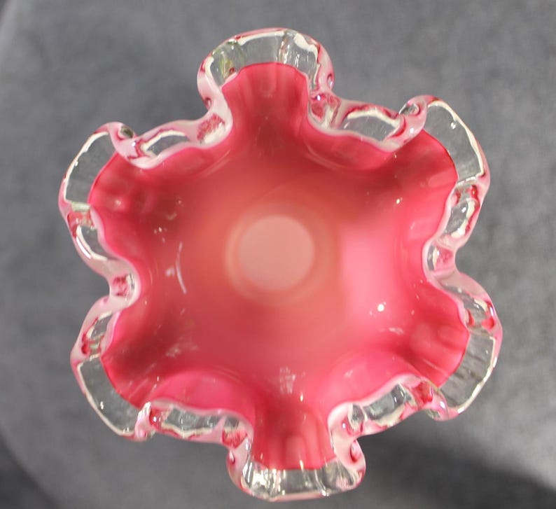 May include: A top-down view of a glass vase featuring a vibrant pink interior that transitions to a lighter, translucent center. The rim is characterized by a distinct, ruffled, and wavy edge with a clear, transparent glass border.